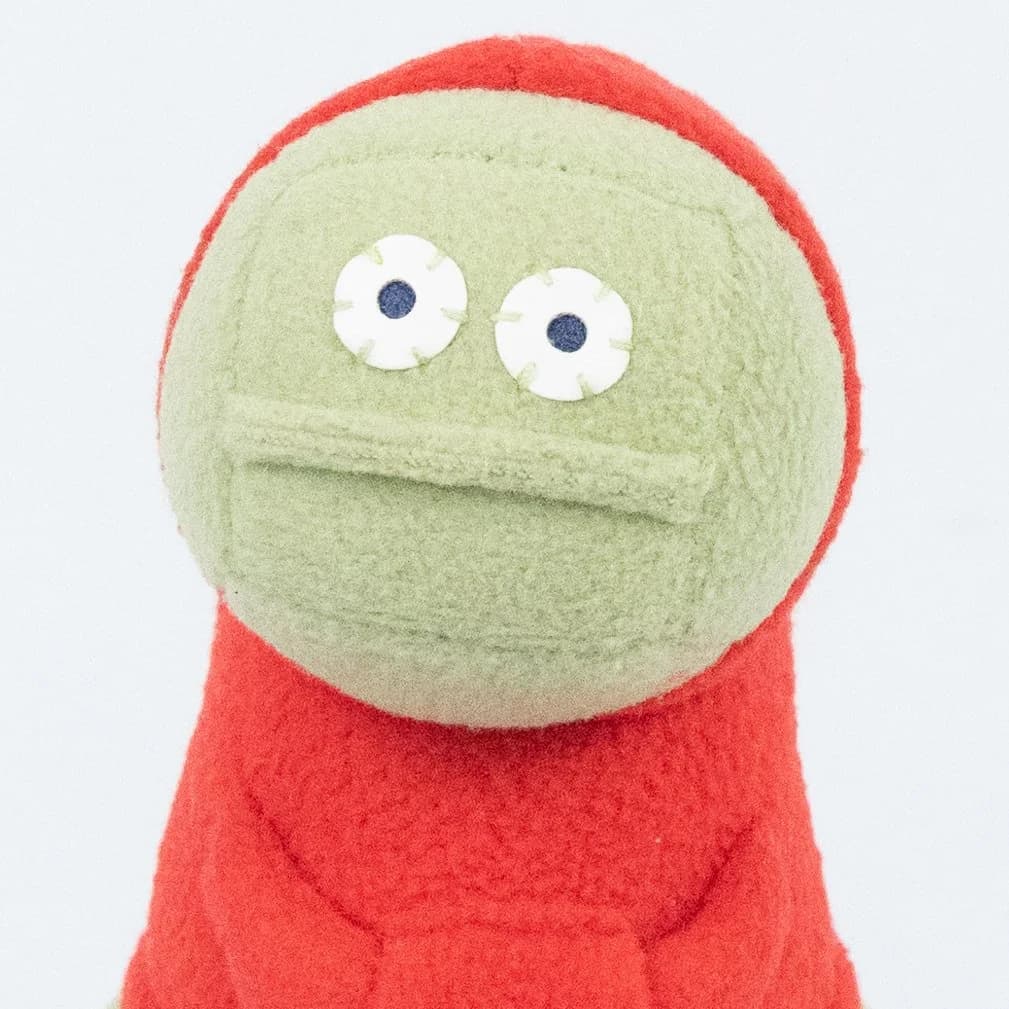 Colin plush, a round green-faced monster character with googly eyes peeking out of a red onesie.