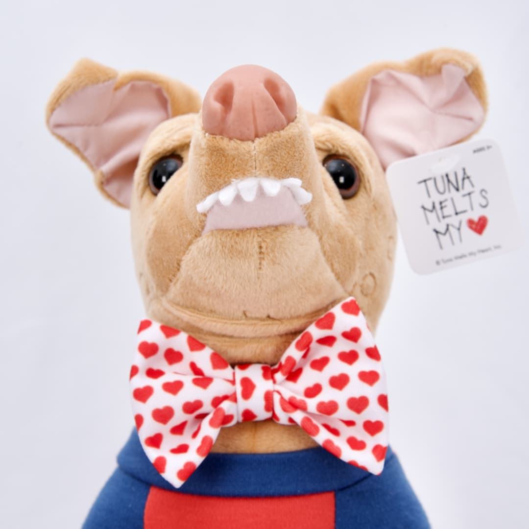 A Perfectly Imperfect Tuna plush with a slightly skewed face and crooked smile, in blue cardigan with red inner shirt and heart bowtie.